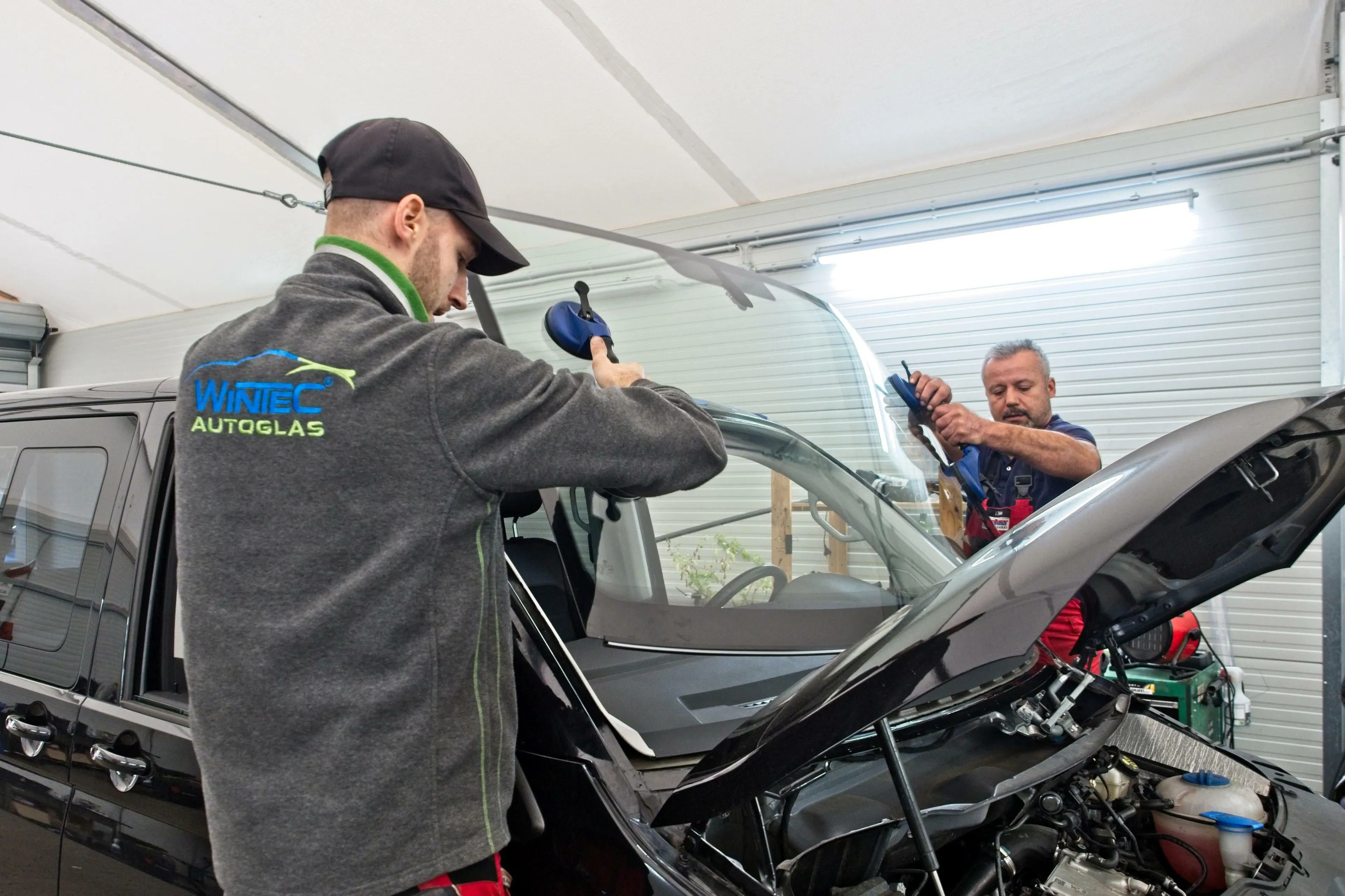 Zwei Automechaniker passen mit Saugnapfwerkzeug eine neue Glasscheibe in vorgesehene Öffnung ein.