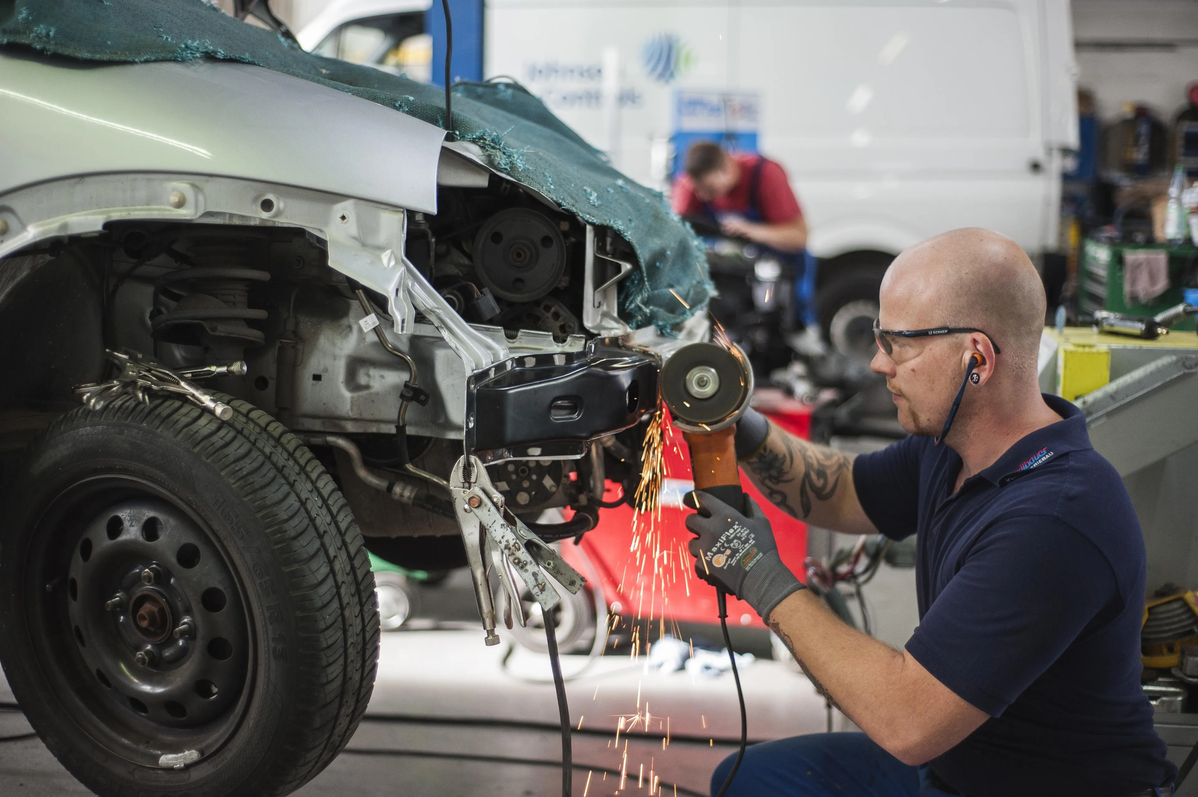 Auf dem Bild ist ein Automechaniker zu sehen, der mit einem Trennschleifer beschädigte Karosserieteile aus dem Auto mit Funkenflug schneidet, um diese durch Neuteile zu ersetzen.